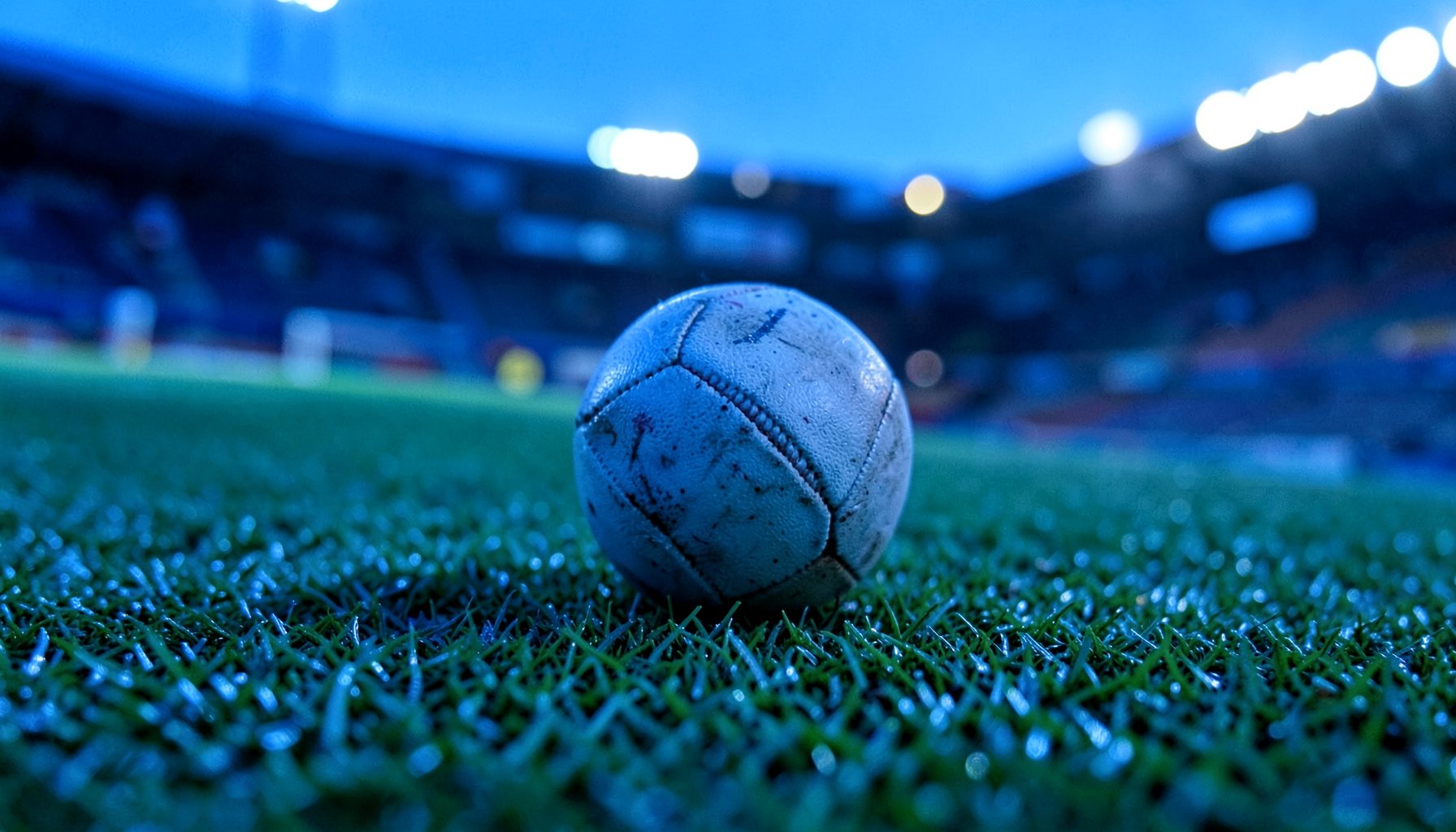 Close-up photograph of a football pitch with a lone ball resting on the grass, stadium floodlights glowing softly in the misty background