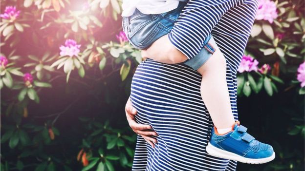 Close-up of a pregnant woman's belly with a child's legs wrapped around it, striped shirt, floral green background