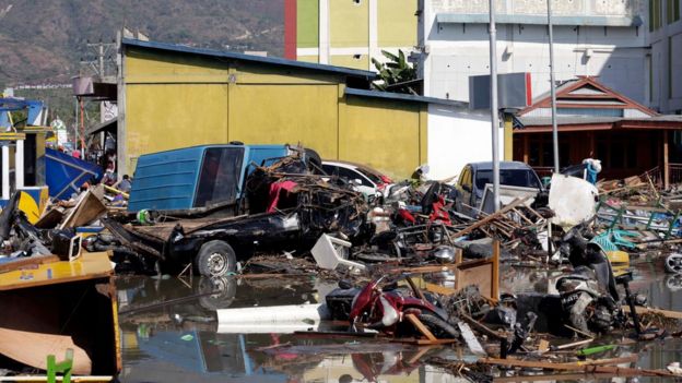 Aftermath of a disaster scene with scattered debris, damaged vehicles, and wreckage near buildings under overcast skies