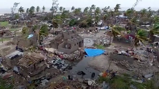 Aerial view of a storm-damaged village with destroyed buildings and bent palm trees along a coastline