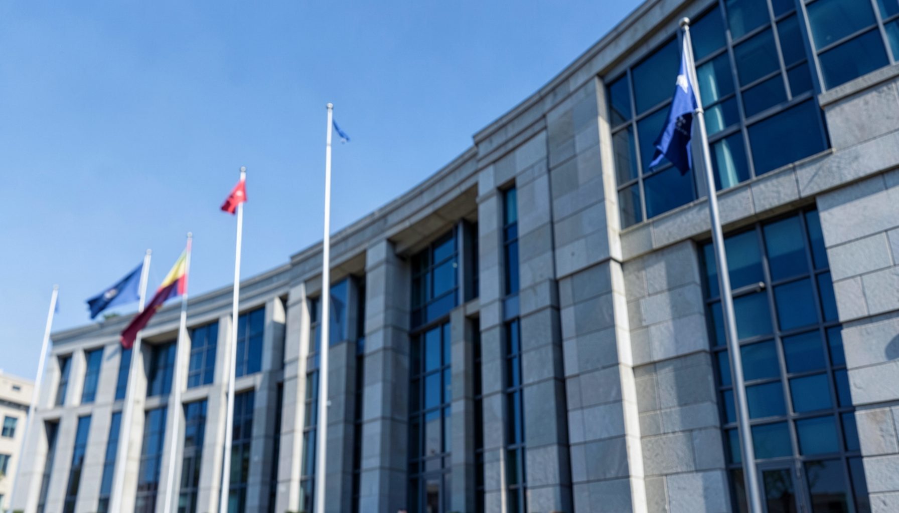A formal institutional building facade with flags, clear sky, stone and glass architecture in neutral grey tones